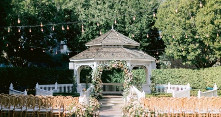 An outdoor ceremony setup with a gazebo, chairs, a flower-lined aisle and string lights overhead