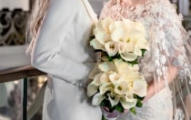 Two women in formalwear hold matching bouquets as they smile and gaze into each other’s eyes