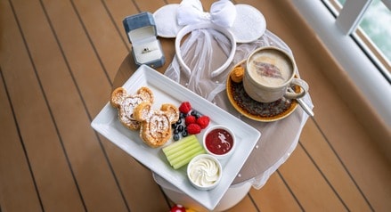 A celebratory breakfast setup on a Disney Cruise Line ship featuring a plate of Mickey Mouse shaped waffles, a coffee cup with latte art on top, a Minnie Mouse headband with a veil and an open ring box