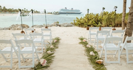 Beachside wedding ceremony setup with palm fronds topped by shells lining the sandy aisle and a Disney cruise ship nearby