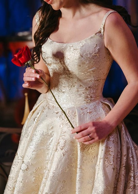 A bride holding a rose while wearing a Disney Fairy Tale Weddings 35th Anniversary Collection Belle inspired wedding gown featuring embroidery, an A line silhouette and a scoop neckline