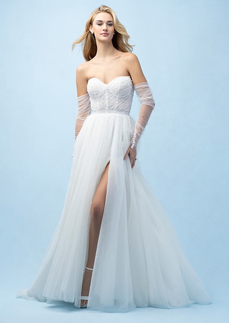 The front of a bride wearing an Ariel inspired A line gown featuring a sweetheart neckline, lightly beaded fabric, a high slit in the skirt and sheer lower arm wraps The front of a bride wearing an Ariel inspired A line gown featuring a sweetheart neckline, lightly beaded fabric, a high slit in the skirt and sheer lower arm wraps