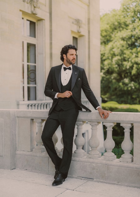 A man in a black tuxedo and bow tie walking across a lawn near a stone archway