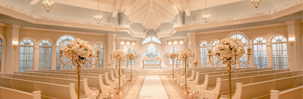 Sunlight shines through the arched stained glass windows of a wedding chapel with chandeliers and a high beam ceiling
