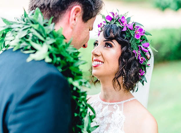 A man and woman, wearing a traditional Hawaiian wedding lei and head band, gaze into each other’s eyes