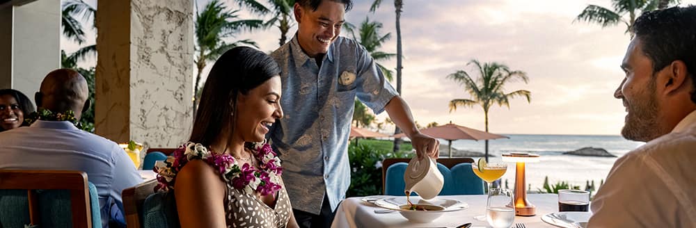 A Cast member serving a couple at an outdoor restaurant with an ocean view and palm trees