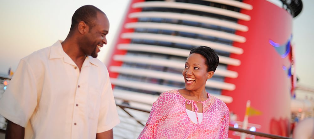 A man and a woman smile in front of the funnel of their Disney Cruise Ship  
