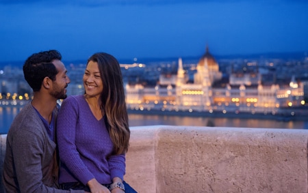 A man and a woman cuddle on a balcony at dusk with the city in the background 