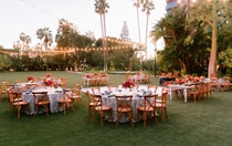 An outdoor wedding reception area with 9 set tables under string lights surrounded by palm trees