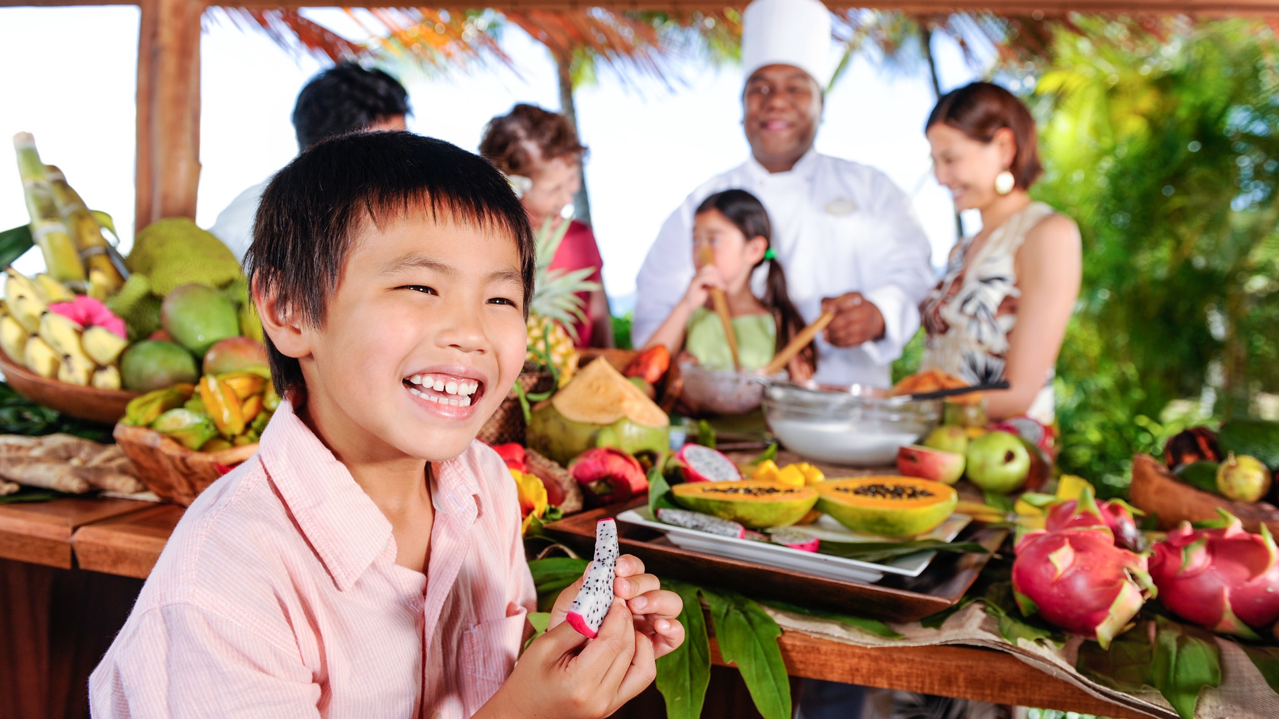 A little boy eating fruit while a chef teaches his family to cook