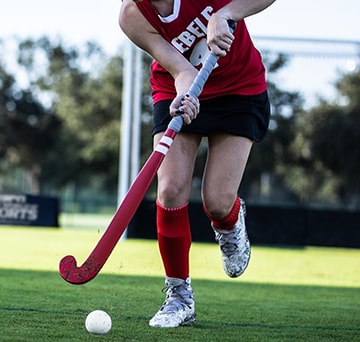 A girl in a Rebels field hockey uniform controls a ball with her stick as she runs down field
