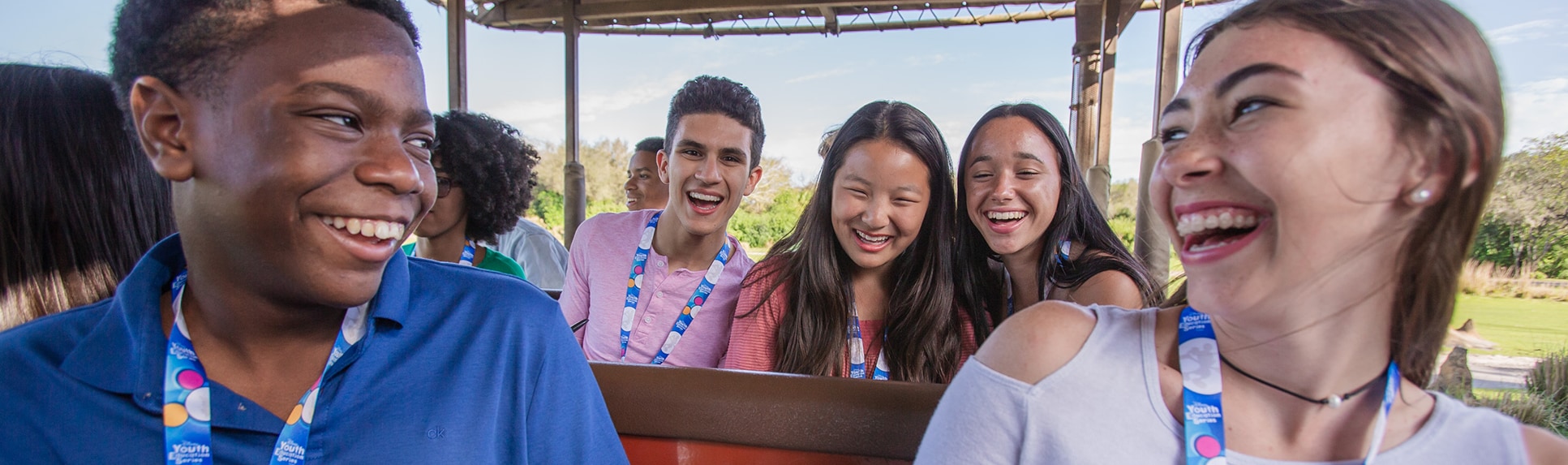 A group of teenagers in an open-air vehicle smiling at each other as they ride through a savanna